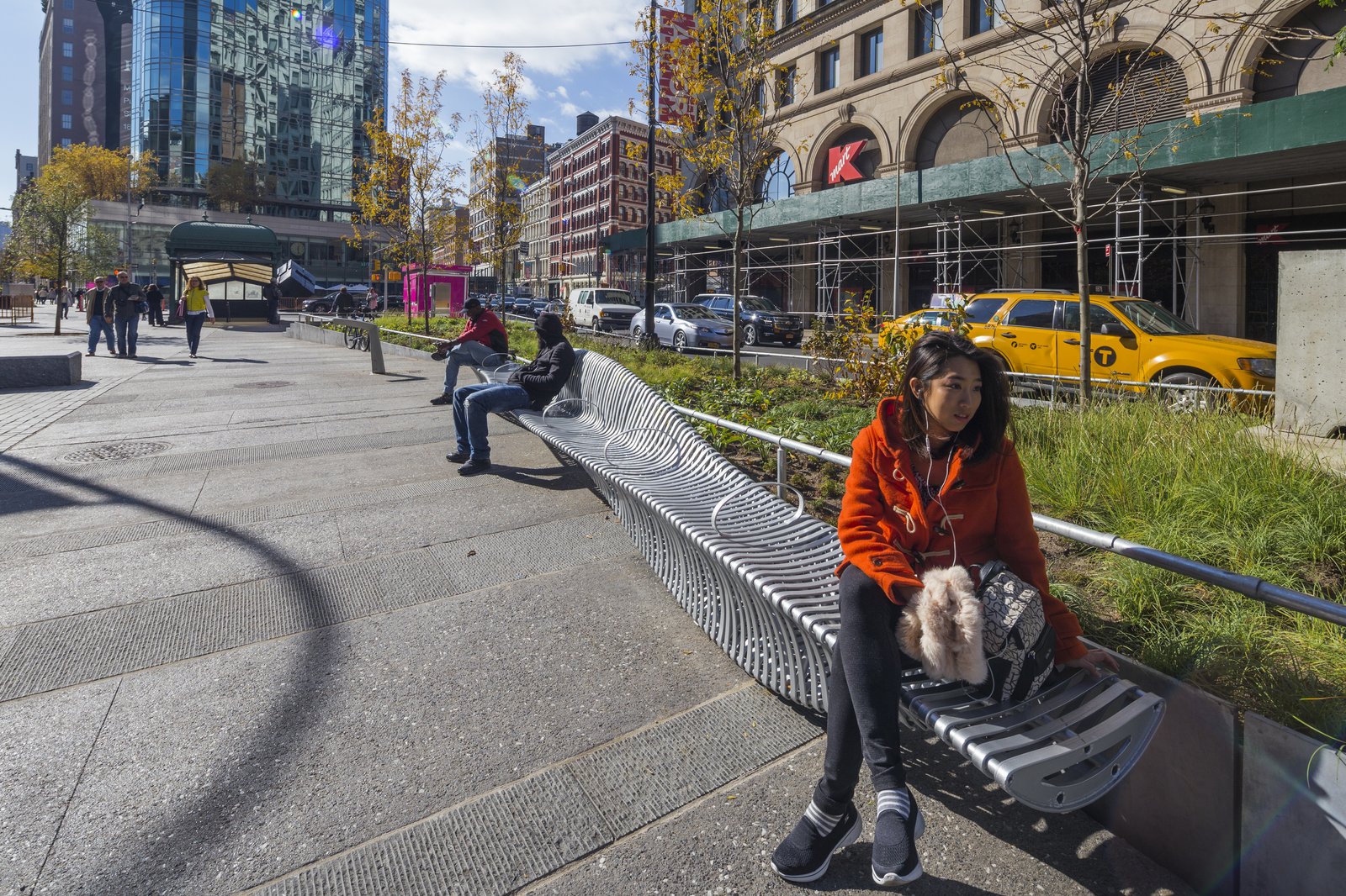 People seated on a zipper bench with trees, buildings, and yellow taxis in the background.