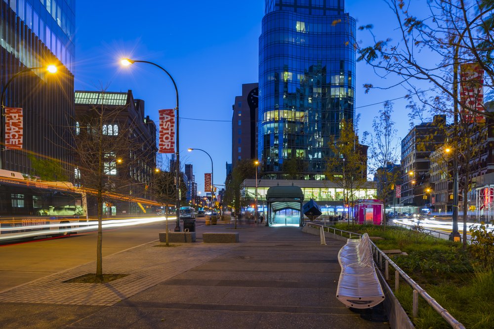 Evening view of Astor Place with illuminated buildings, a subway kiosk, and a metal bench.