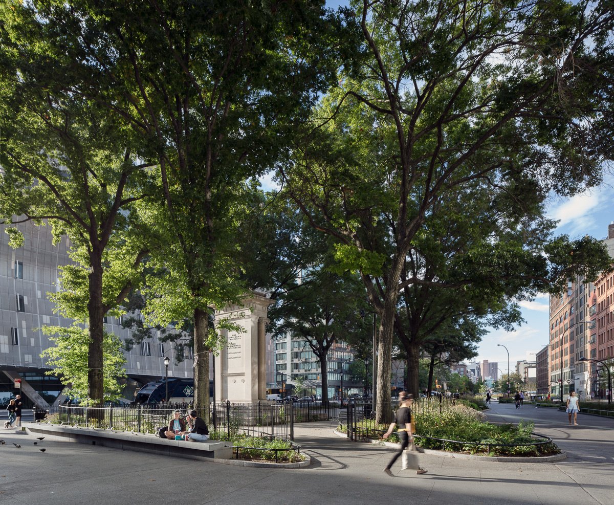 Tree-lined plaza with benches, greenery, and a historic monument, framed by urban buildings.