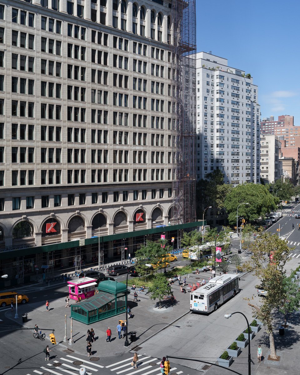 View of a busy urban plaza with a bus, pedestrians, trees, and a historic building facade.