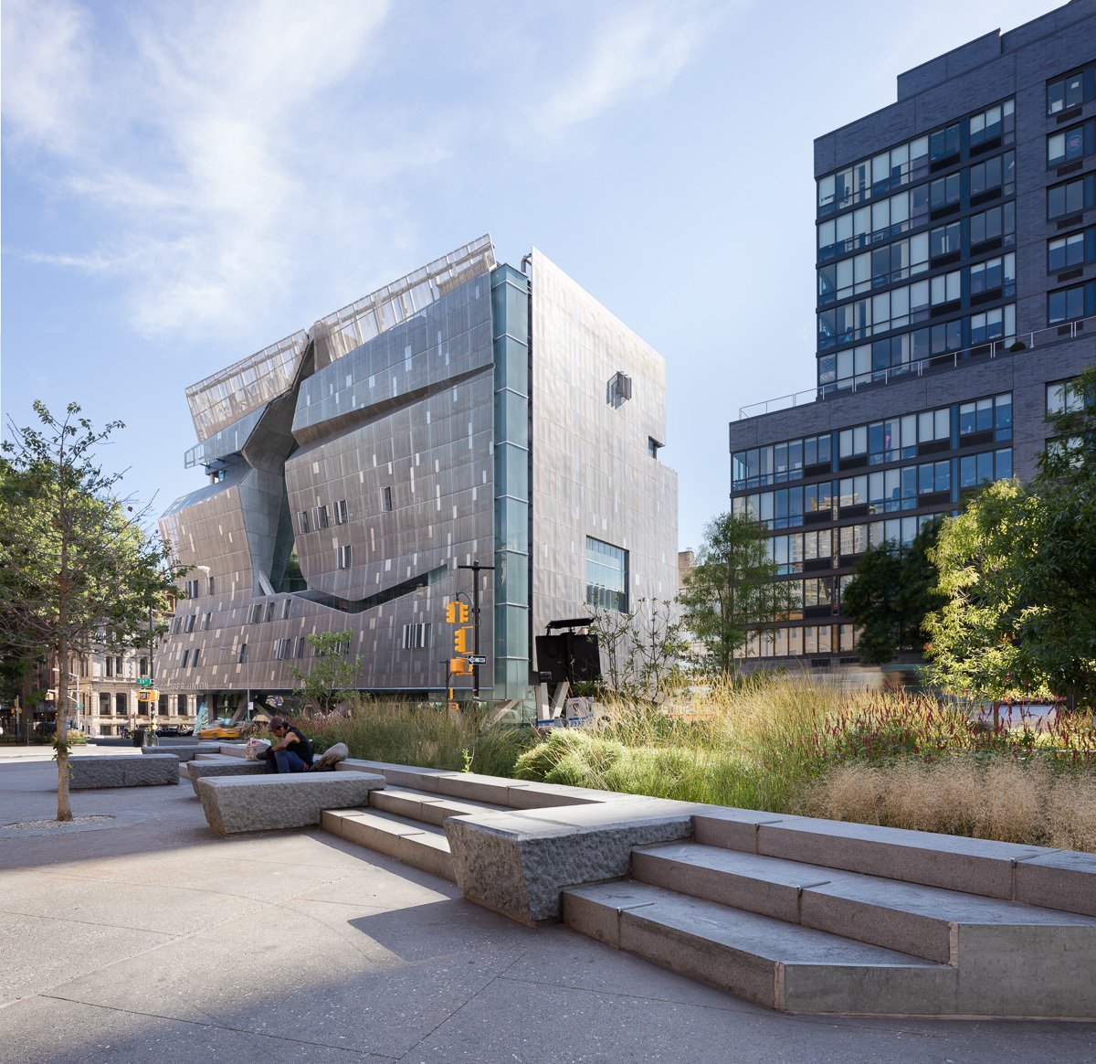 Angular modern building framed by stone seating, steps, and lush plantings in a public plaza.