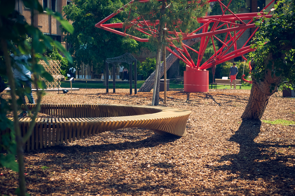 Curved bench under trees with a red structure in the background.