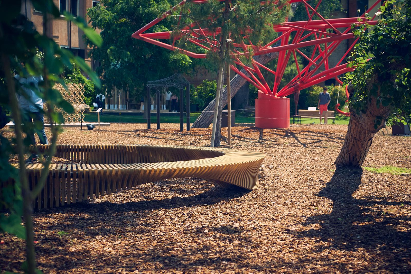 Curved bench under trees with a red structure in the background.