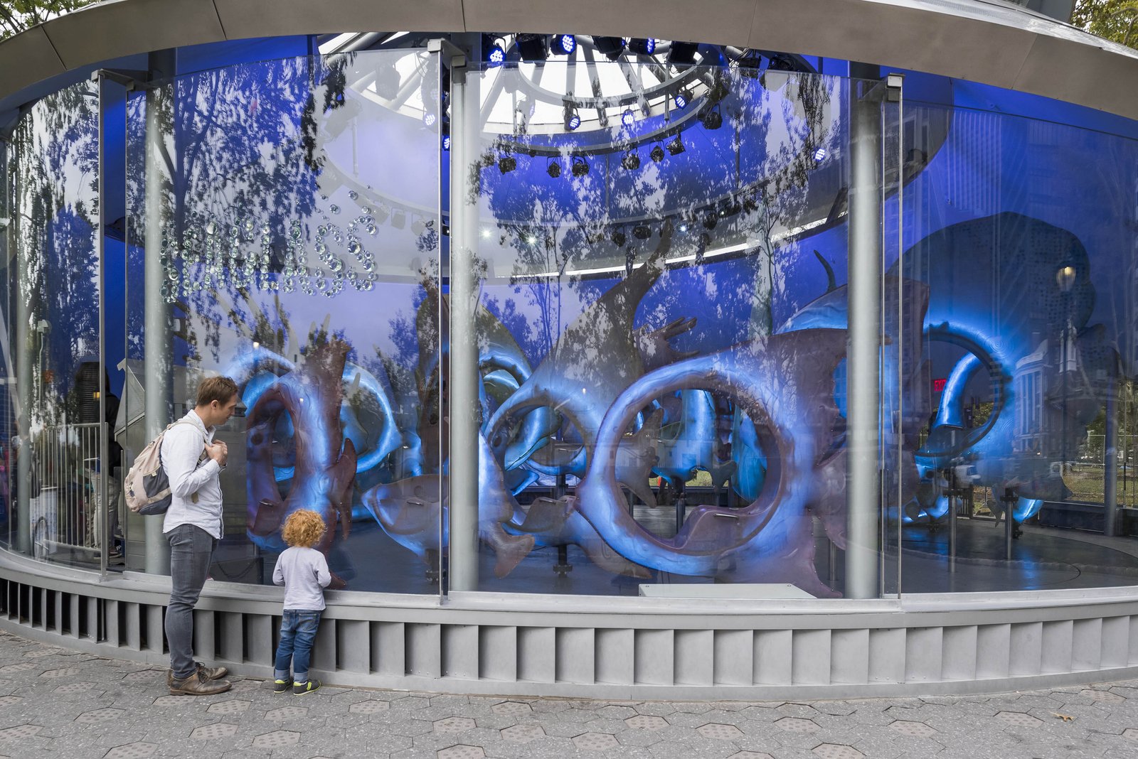 A father and young child stand outside the SeaGlass Carousel, peering through large glass windows at the illuminated fish seats inside.