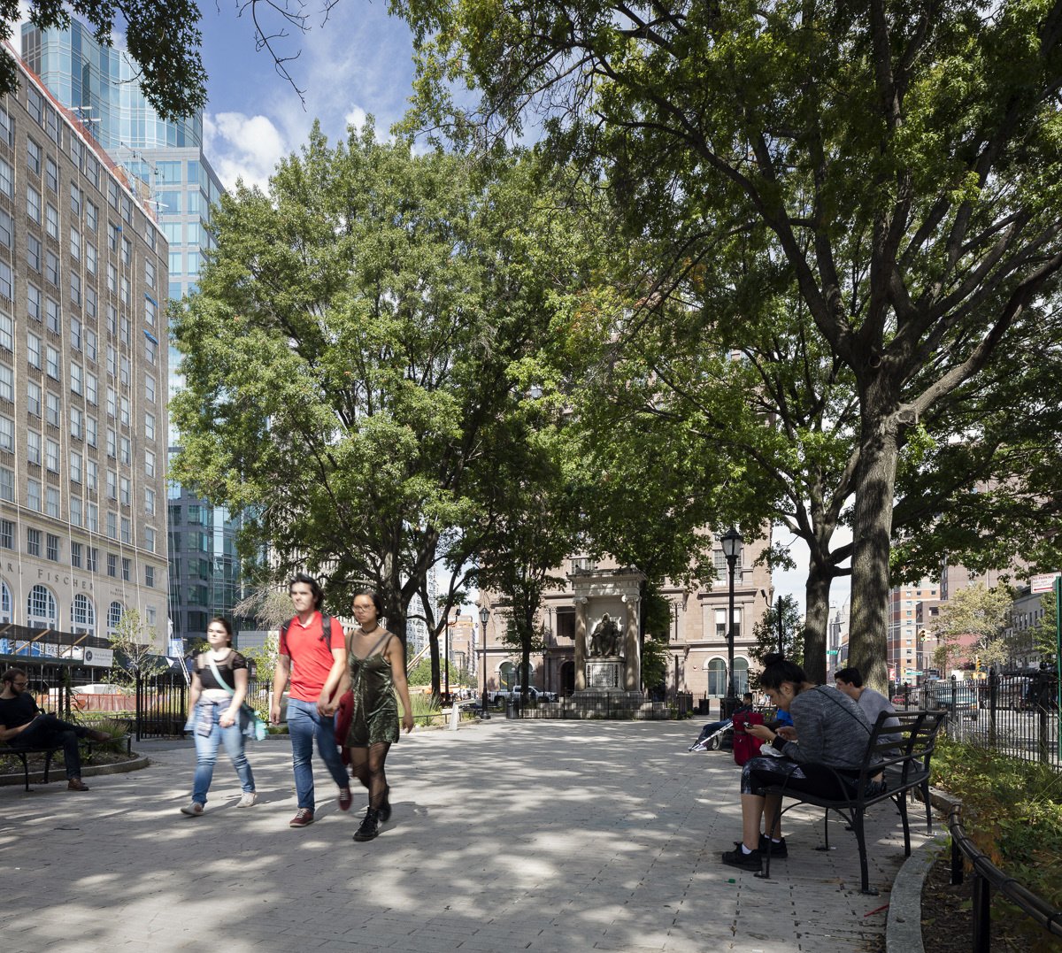 Tree-lined plaza with people walking and sitting on benches, framed by urban buildings and a statue.