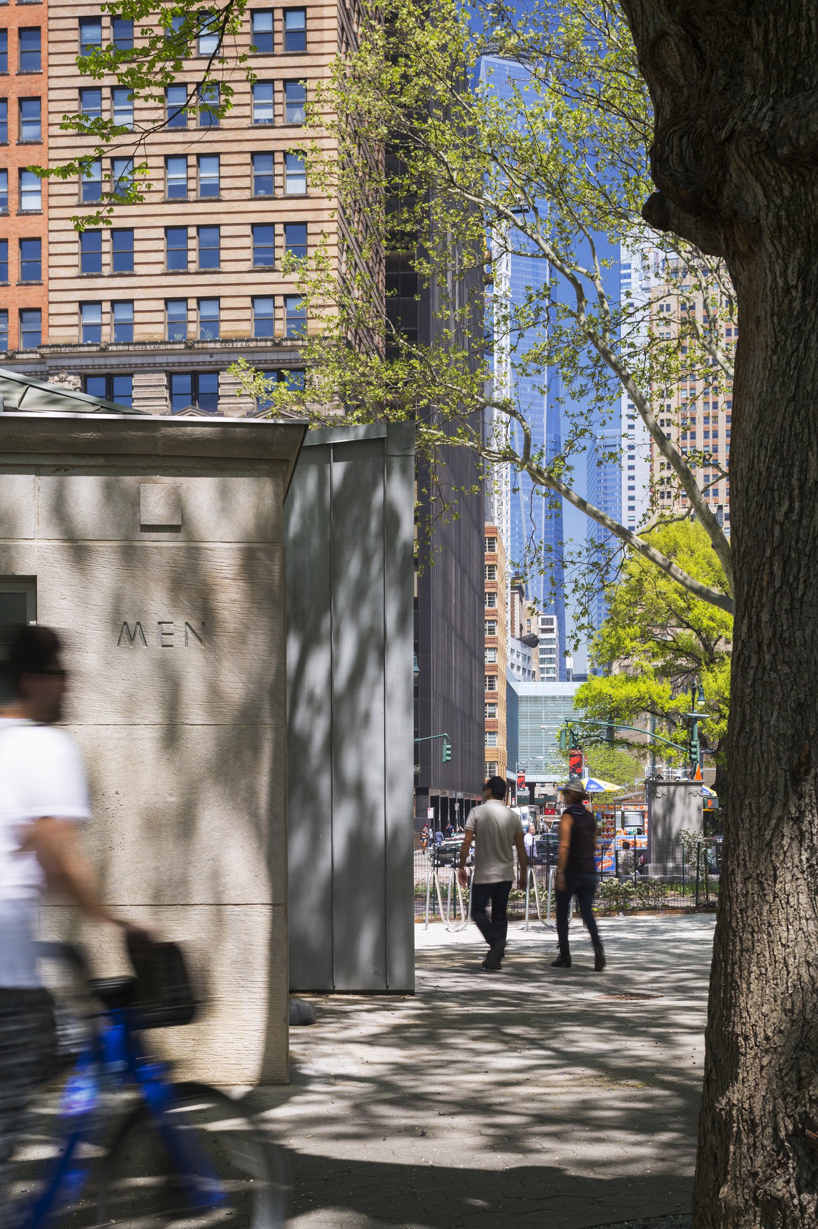 Close-up view of the comfort station corner, framed by trees, with urban buildings in the background.