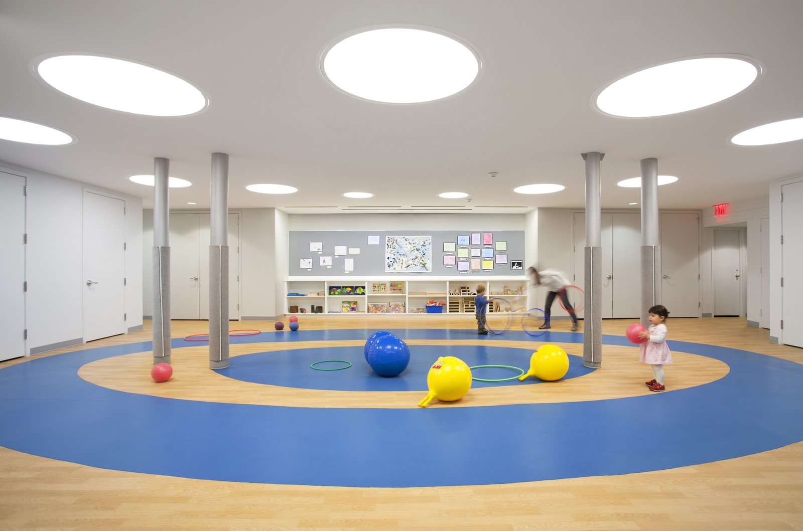 A circular play area with blue flooring, support columns, and children playing with balls and hoops.