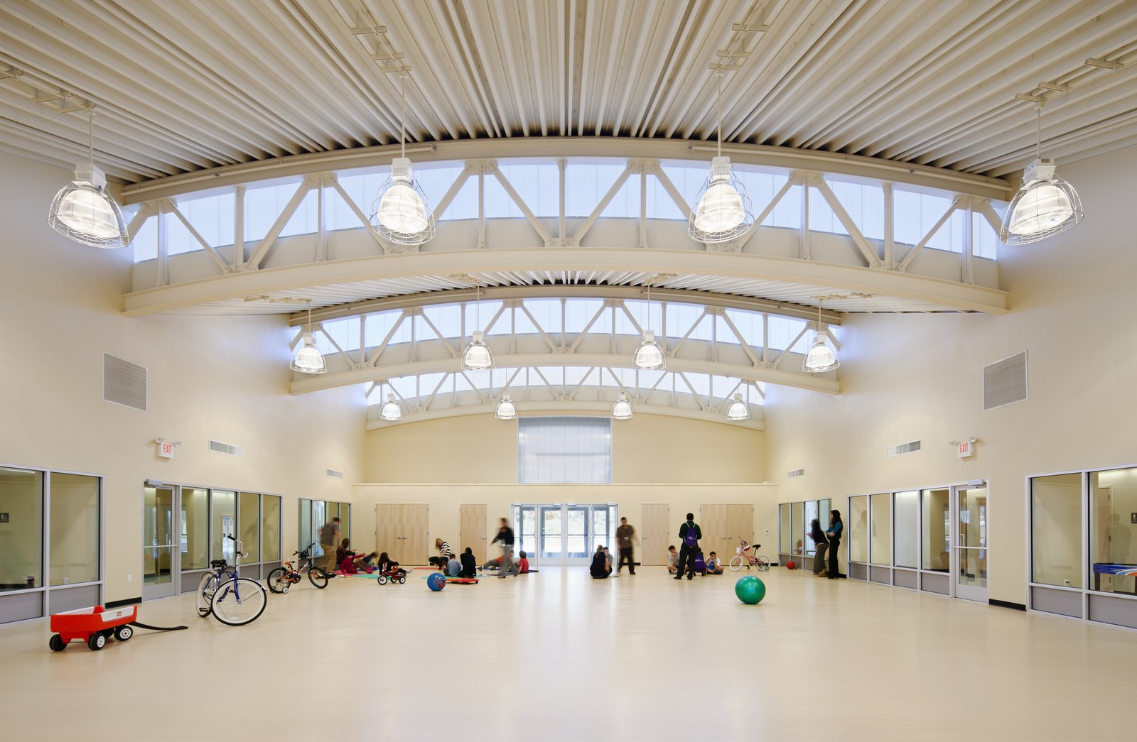 A spacious indoor activity room with arched trusses, clerestory windows, and children playing.