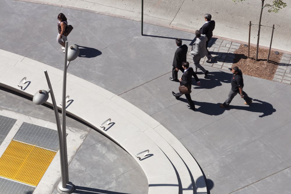 Curved concrete steps with seating, tables, and trees in a shaded urban plaza.