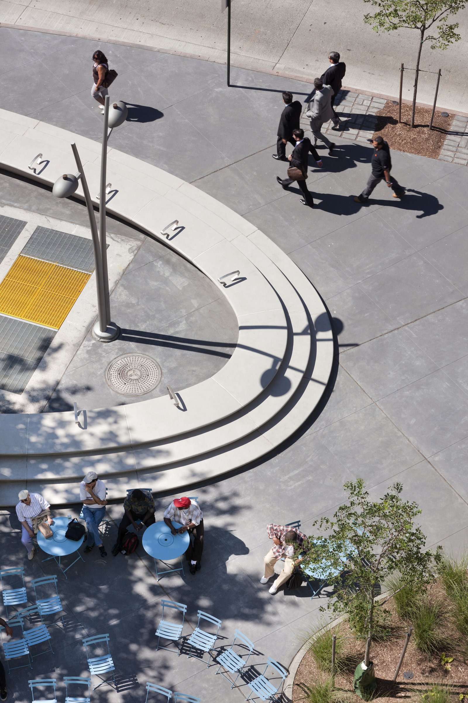 Curved concrete steps with seating, tables, and trees in a shaded urban plaza.