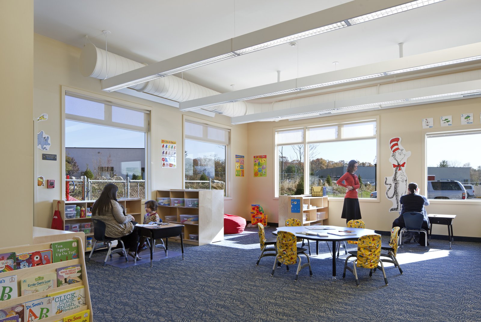 A bright classroom with large windows, child-sized tables, and teachers engaging with students.