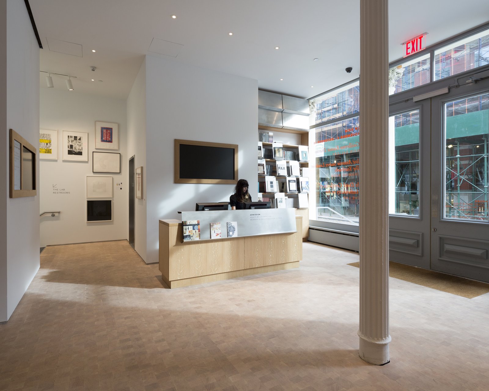 Gallery reception area with light wood desk, white walls displaying artwork, and floor-to-ceiling windows.