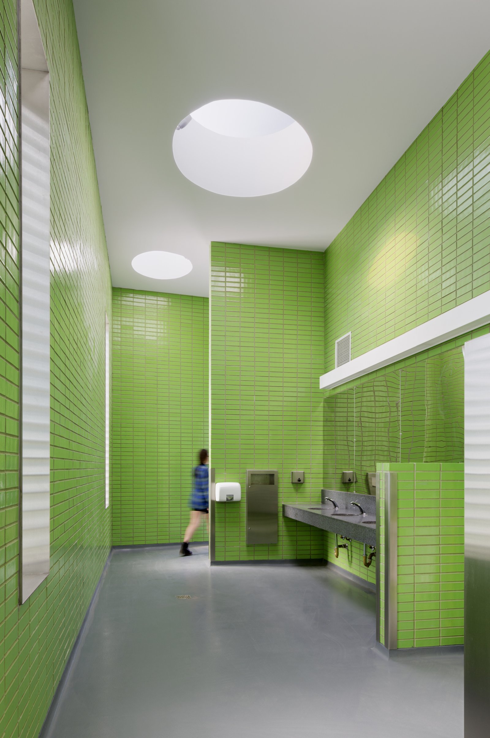 Interior of a restroom with green tiled walls, circular skylights, and stainless steel fixtures.