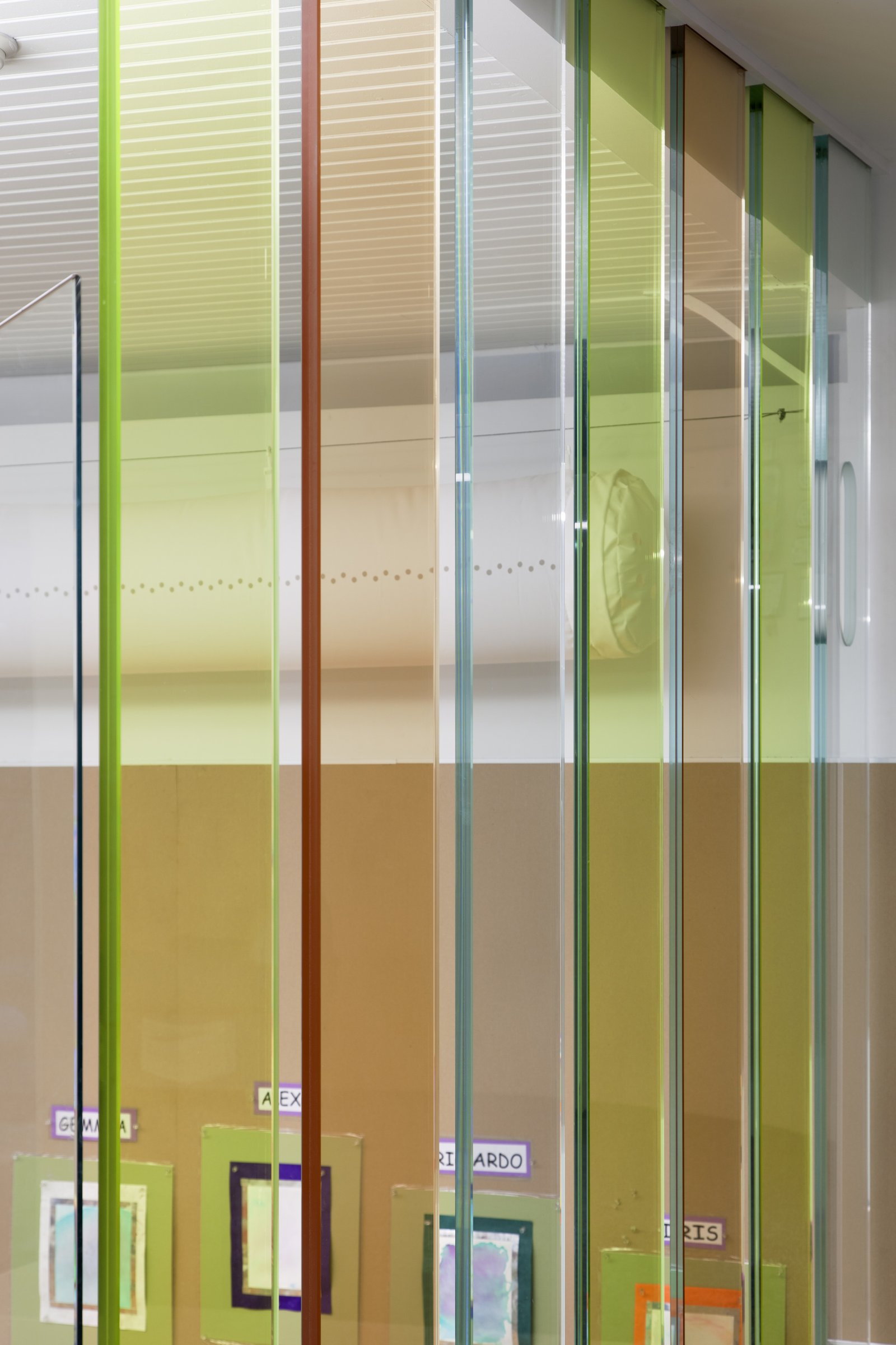 Vertical glass panels in green and amber hues frame a classroom display of children's artwork.