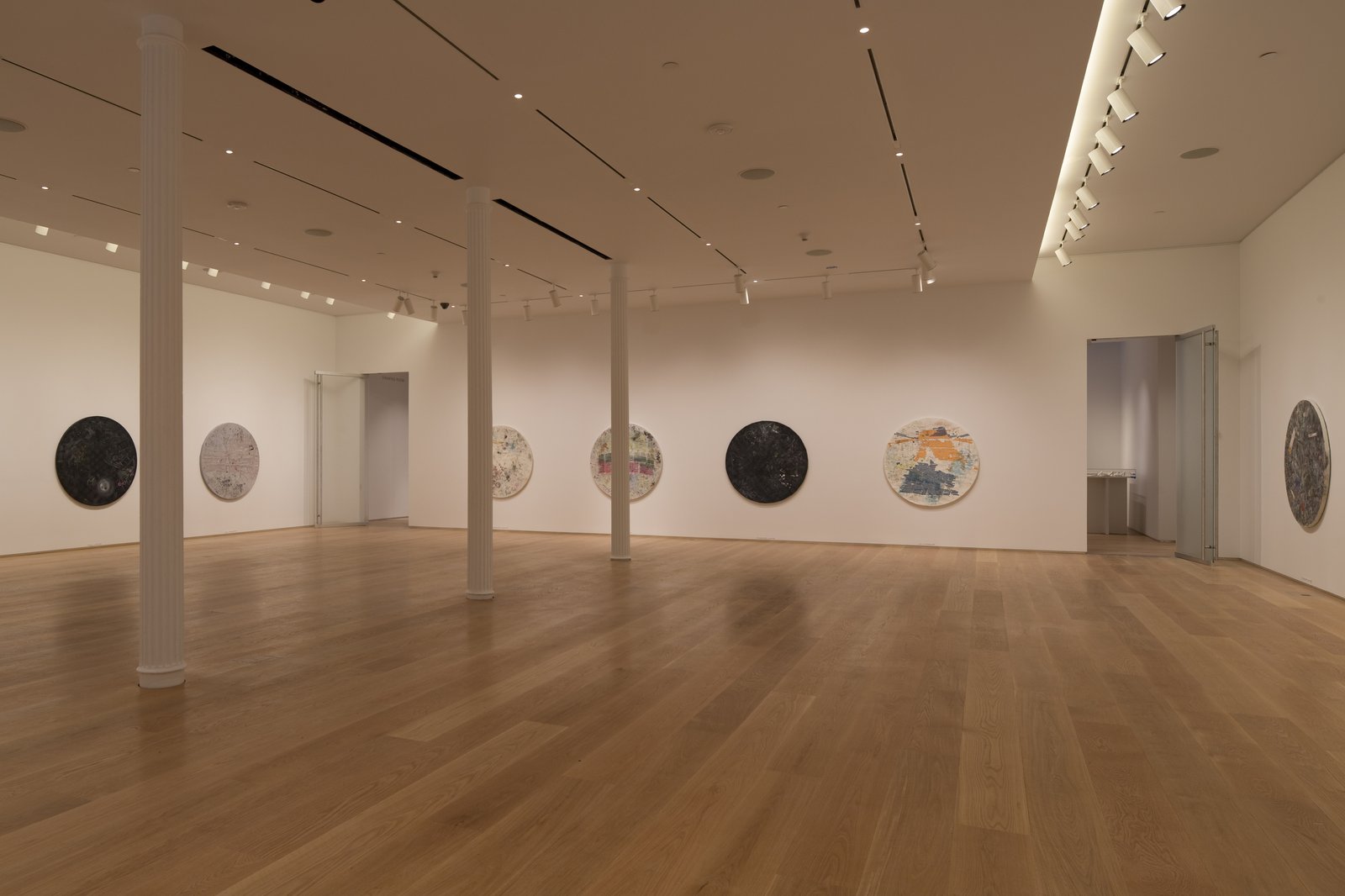 Gallery space with white columns and walls, circular artworks evenly spaced, wide plank oak flooring and track lighting.