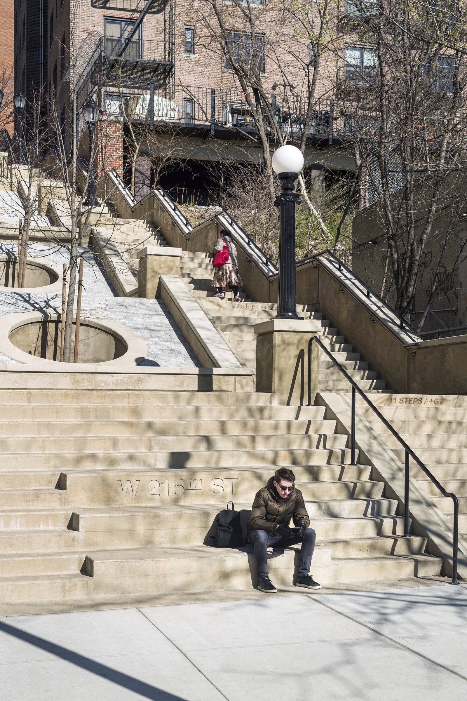 A man sits on engraved concrete steps with handrails and a lamppost; trees and stairs are visible.