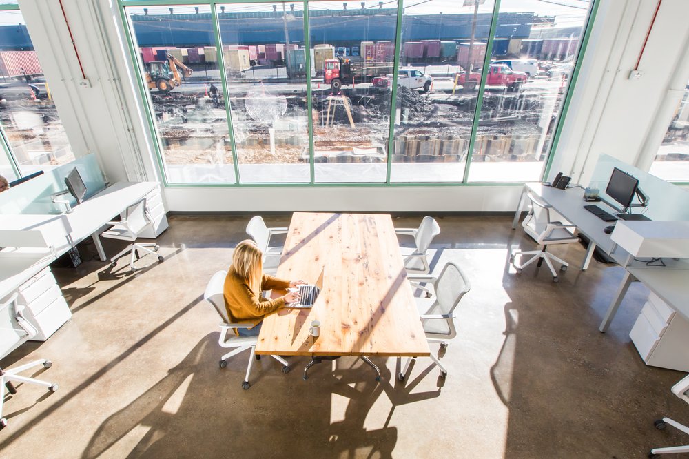 Sunlit office space with large windows overlooking construction, wooden conference table, and white workstations."