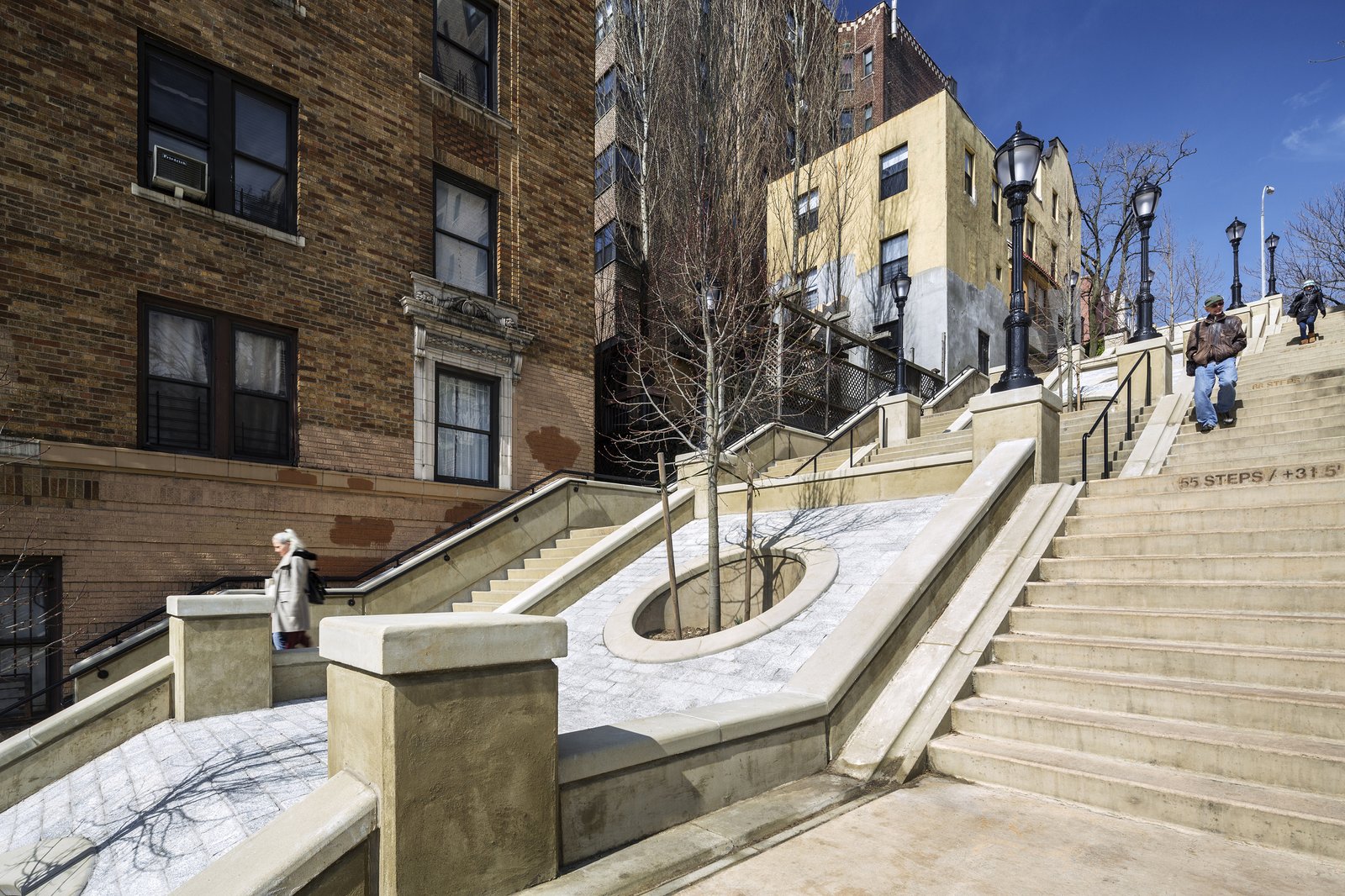 Terraced steps with tree wells, railings, and lampposts between brick and stucco buildings.