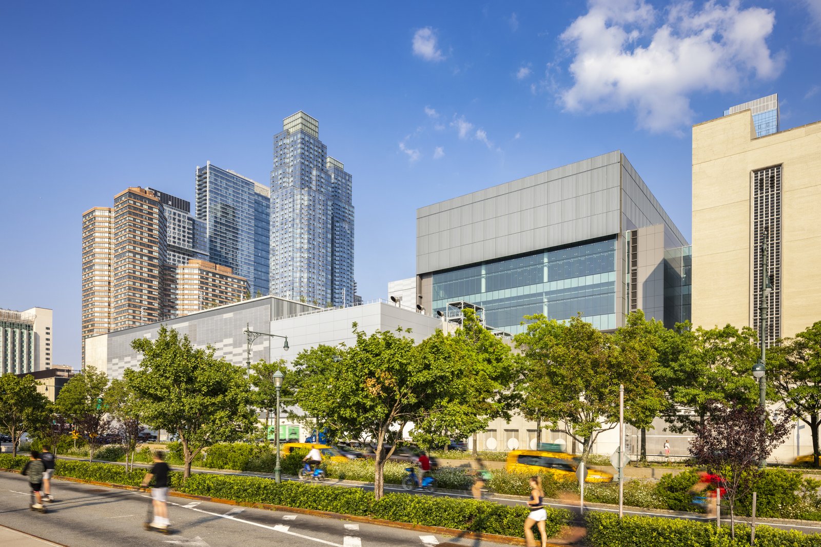 People walk and bike along a tree-lined path in front of a modern building complex, with tall apartment towers behind it.