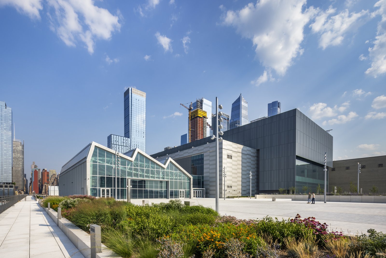 A glass building with a sawtooth roof sits next to a garden filled with colorful plants, while skyscrapers rise behind it.