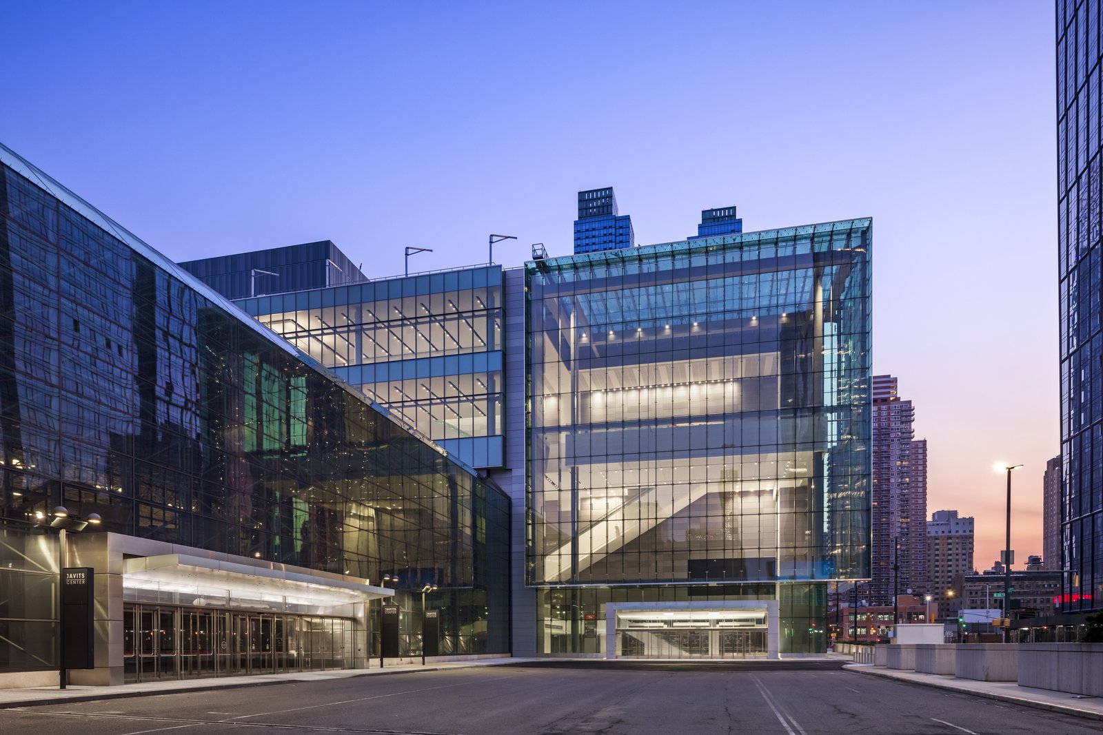 A glass building glows from within at dusk, its transparent walls revealing a bright interior and large staircase inside.