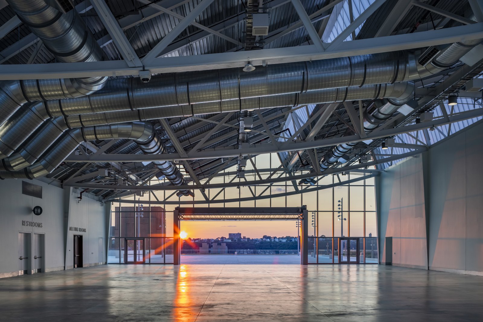 The sunset streams into an empty industrial room through large windows, lighting up exposed ductwork and concrete floors.