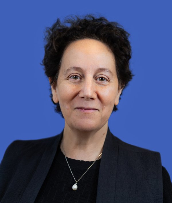 Headshot of Claire Weisz wearing a dark blazer over a black top with a blue background.