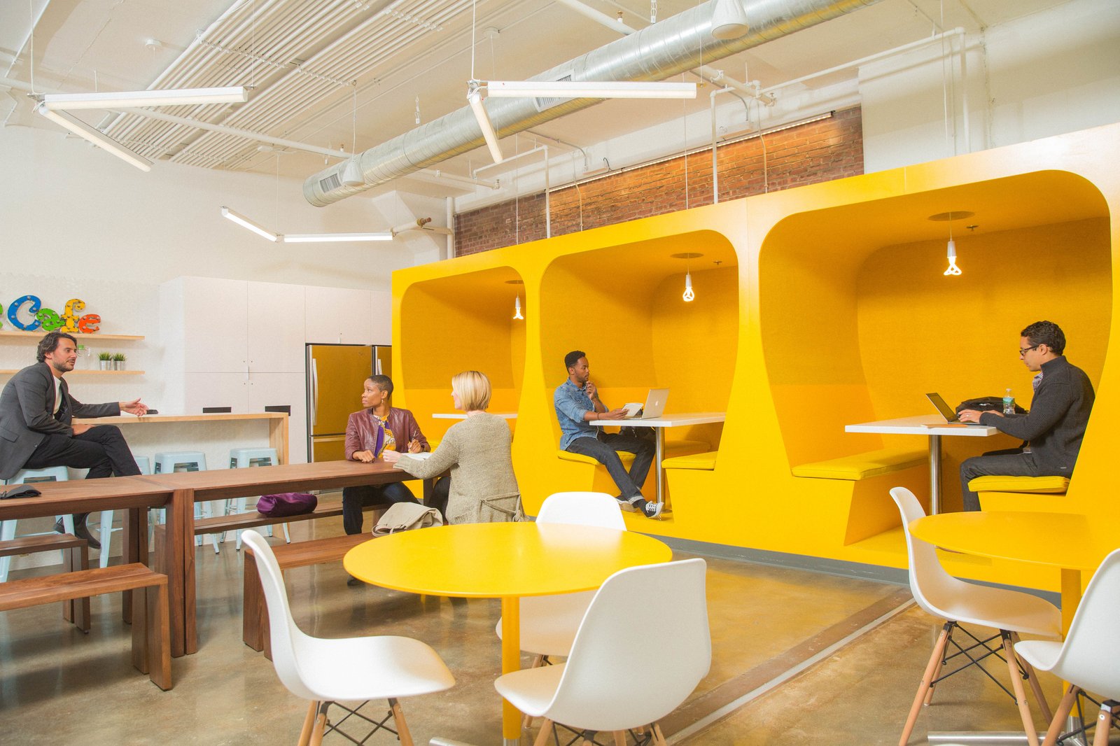 Office break room with bright yellow alcove booths, wooden communal table, and white chairs around tables.