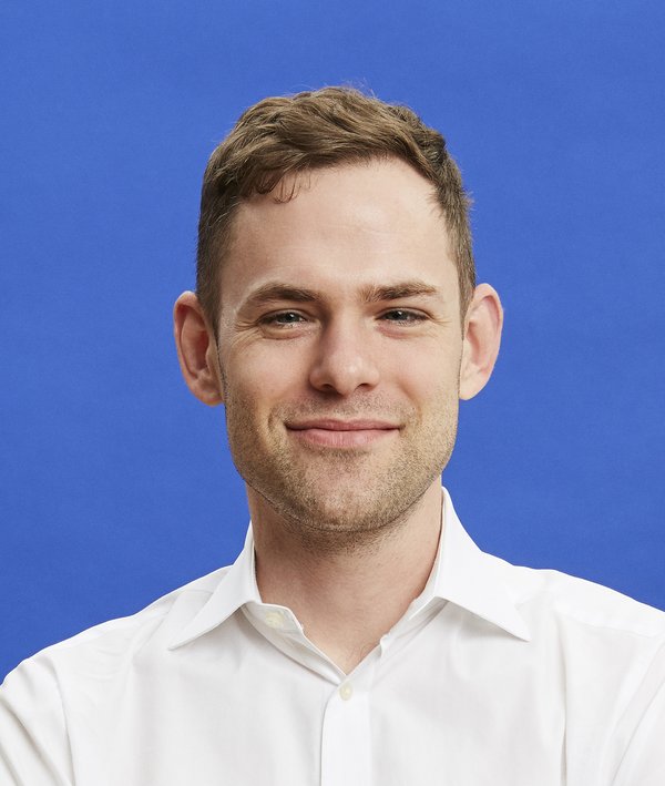 Headshot of Tom Offord in a white shirt against a blue background.