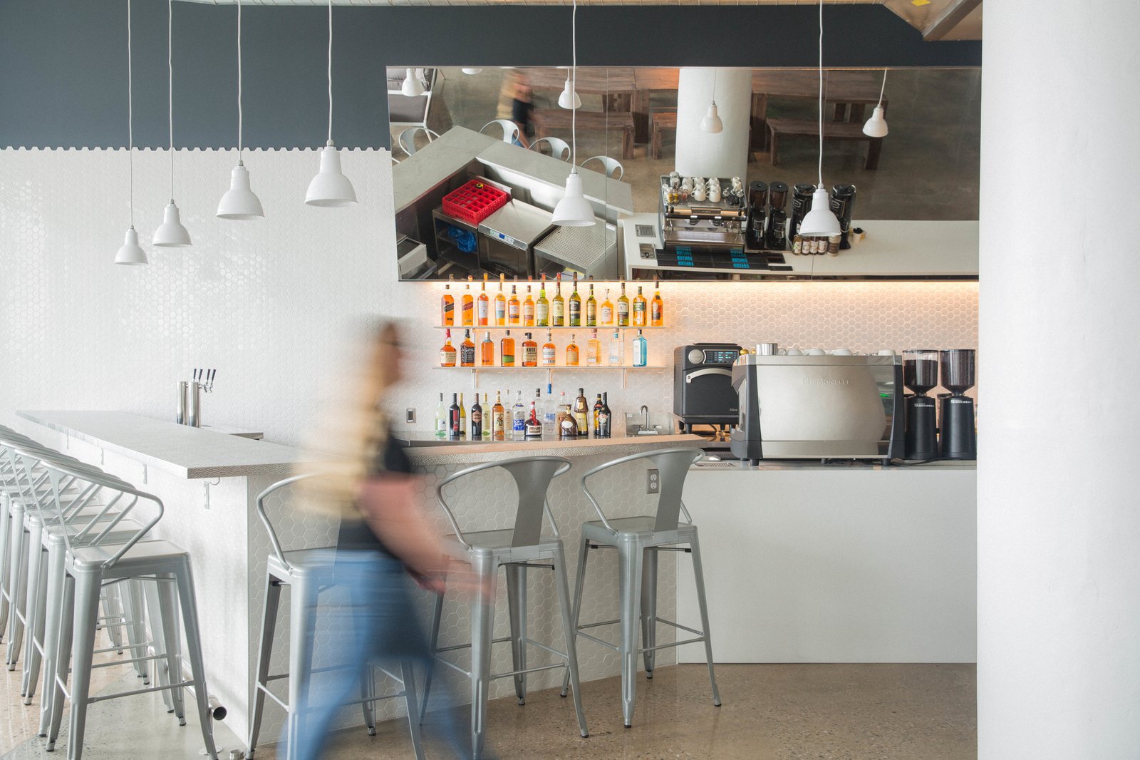 Modern office bar area with metal counter stools, pendant lights, backlit liquor display, and coffee equipment.