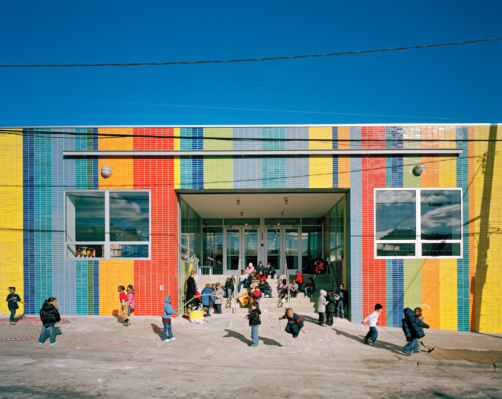 Children play and jump rope in front of the brightly colored school building.