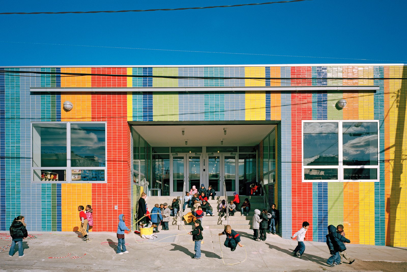 Children play and jump rope in front of the brightly colored school building.