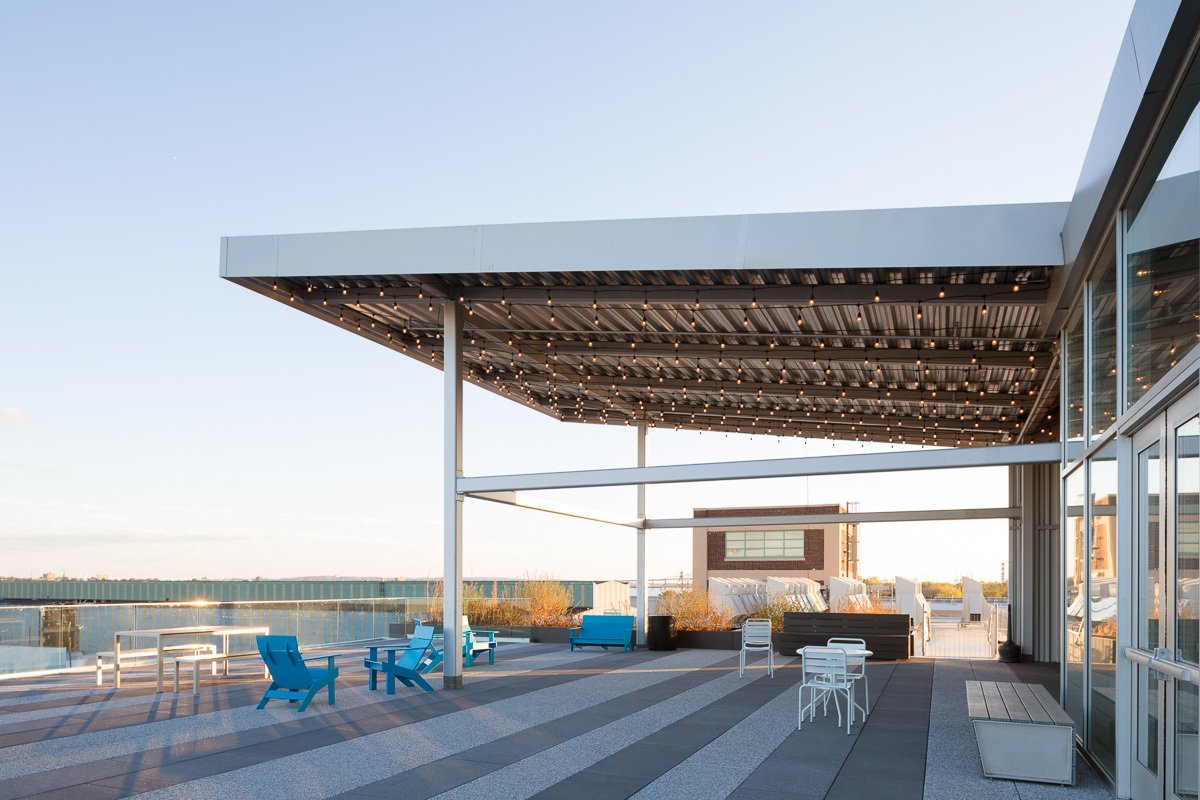 Modern rooftop terrace with striped flooring, string lights overhead, and blue lounge chairs facing city views.