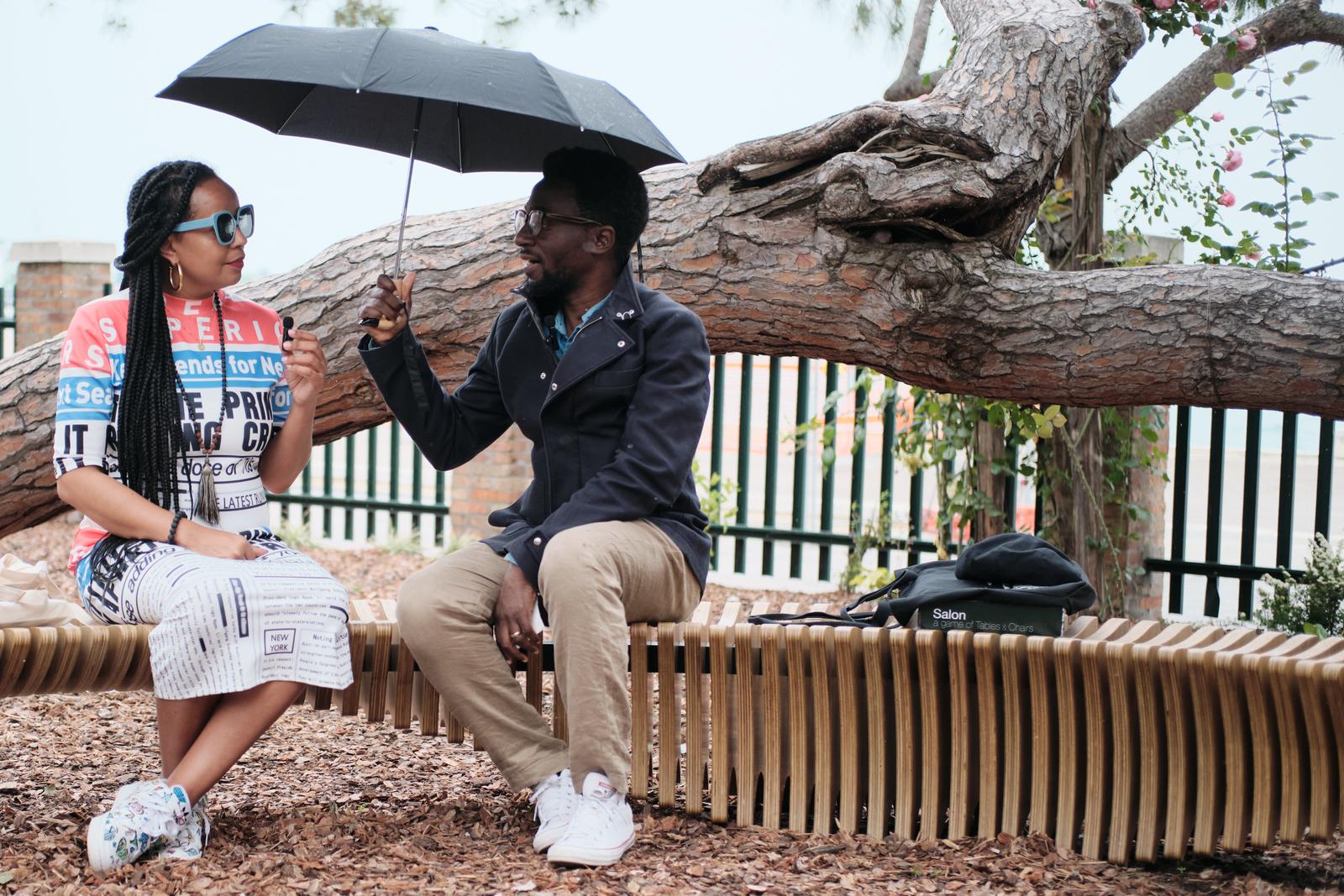 Two people sitting on a wooden bench, sharing an umbrella, under a tree.