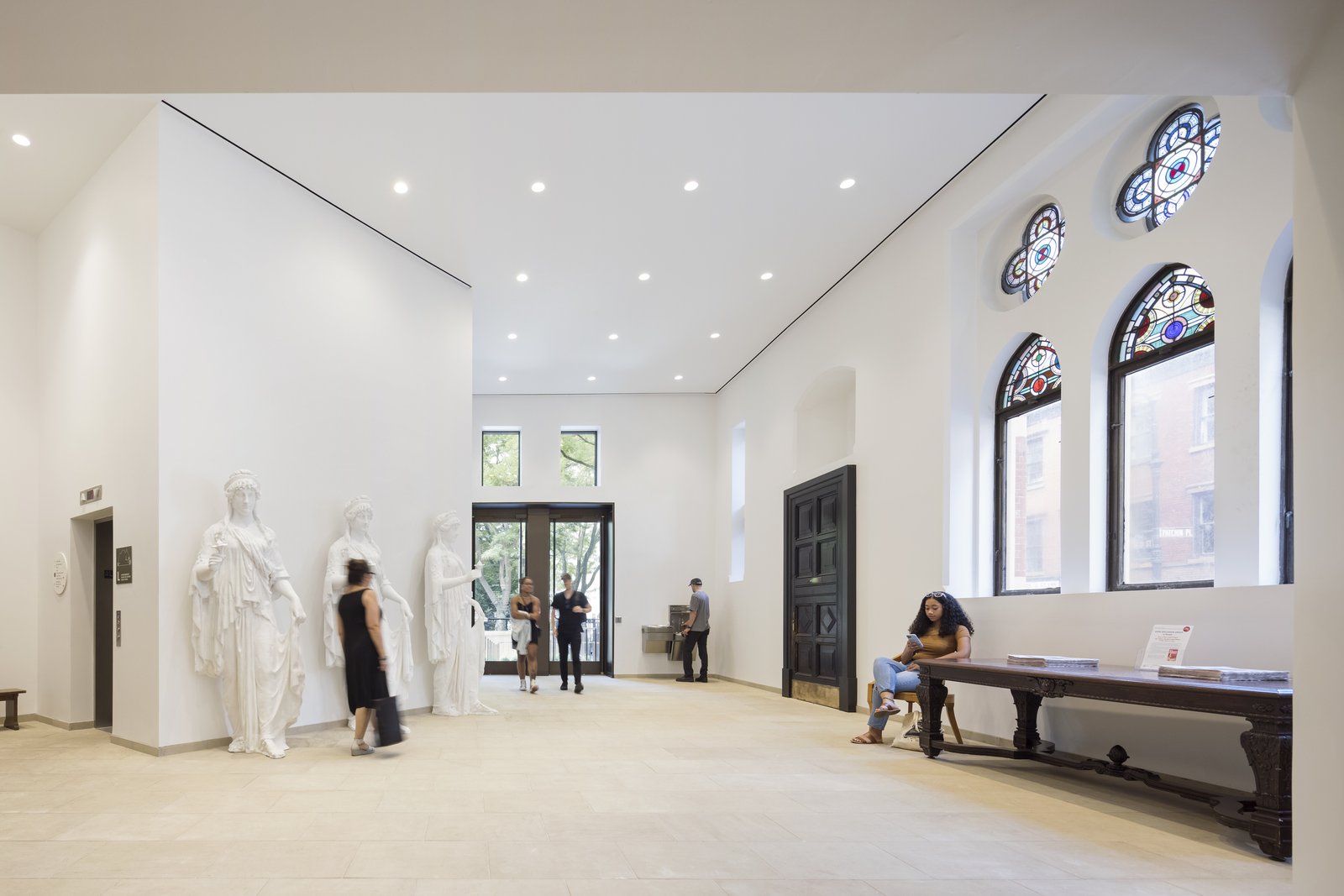 A bright white room features three classical statues, stained glass windows, and a wooden bench along one wall