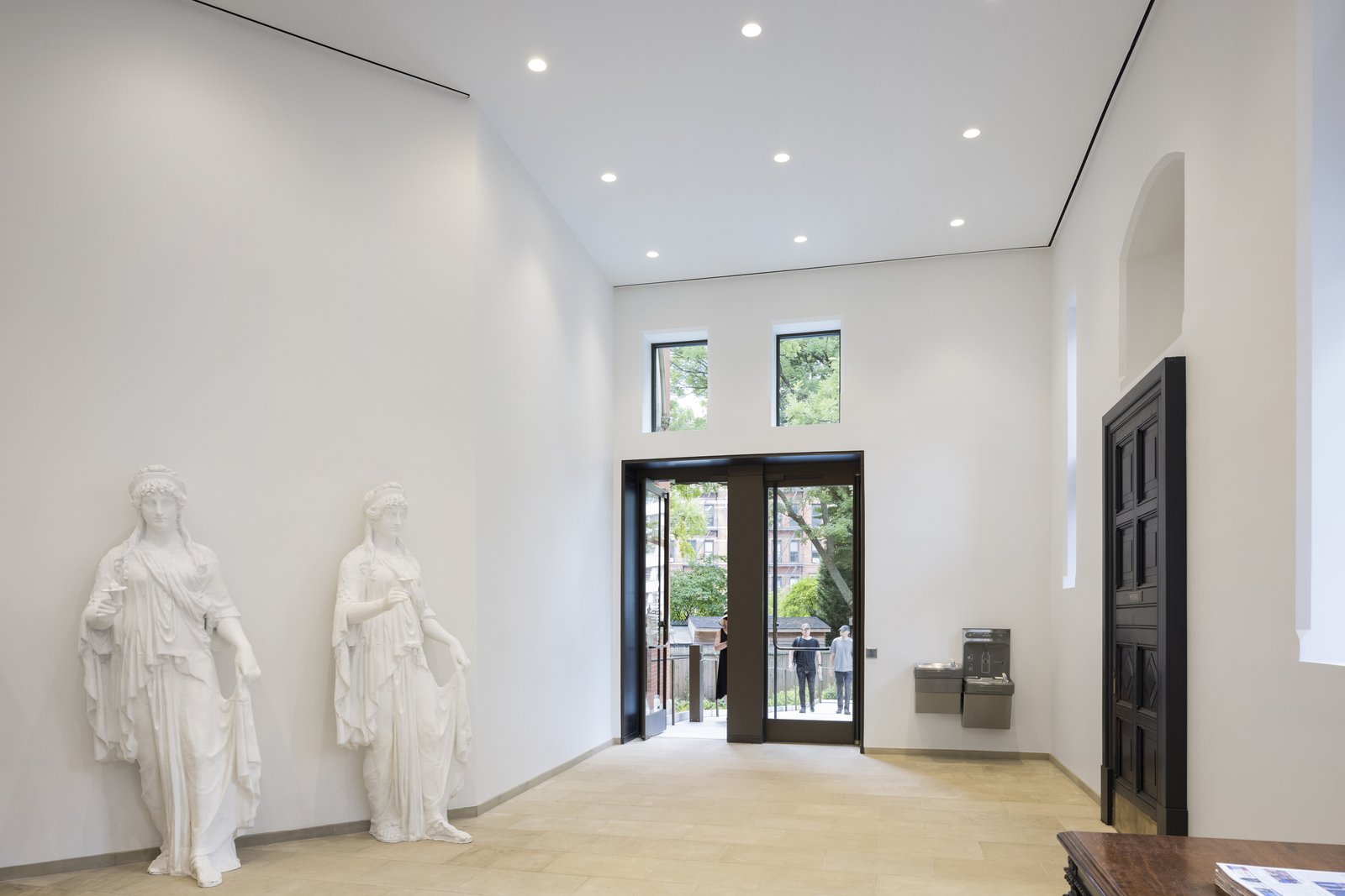 A bright white hallway has two classical statues, glass doors at the far end, and a black wooden door on the side.