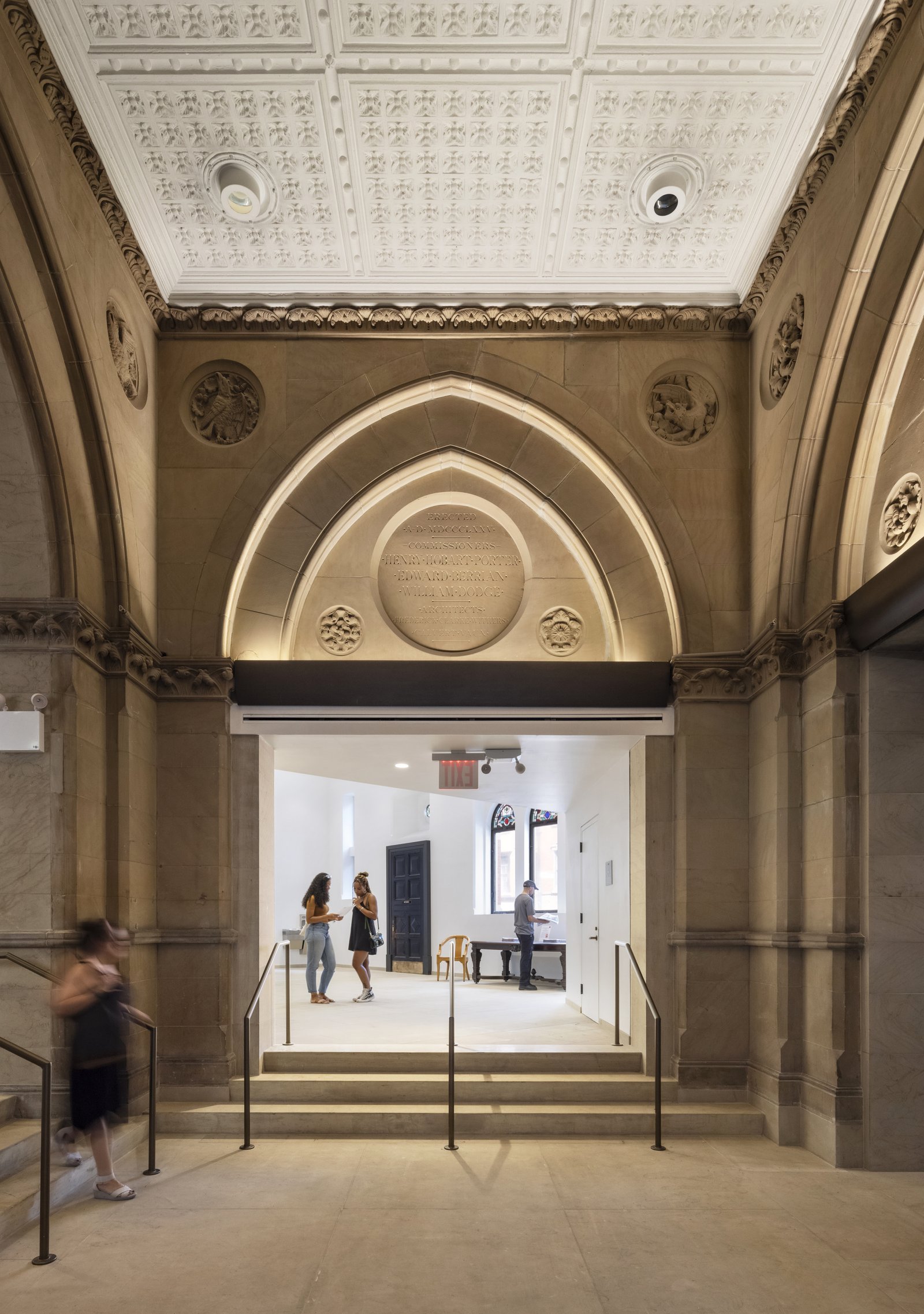 A stone archway leads to a bright room through steps with handrails, while carved details decorate the walls and ceiling.