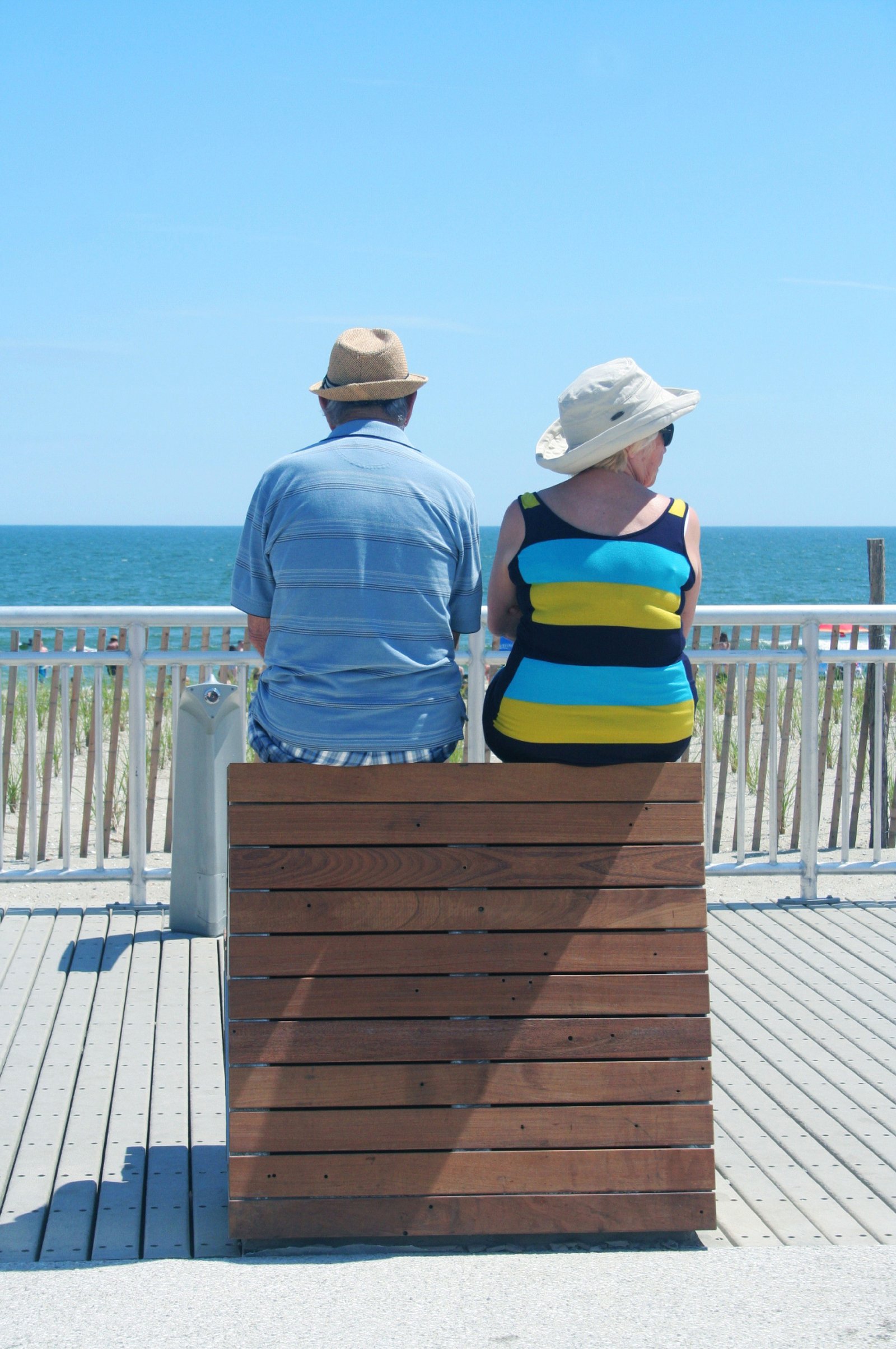 Two people sitting on a wooden bench on the Rockaway Boardwalk, facing the ocean.