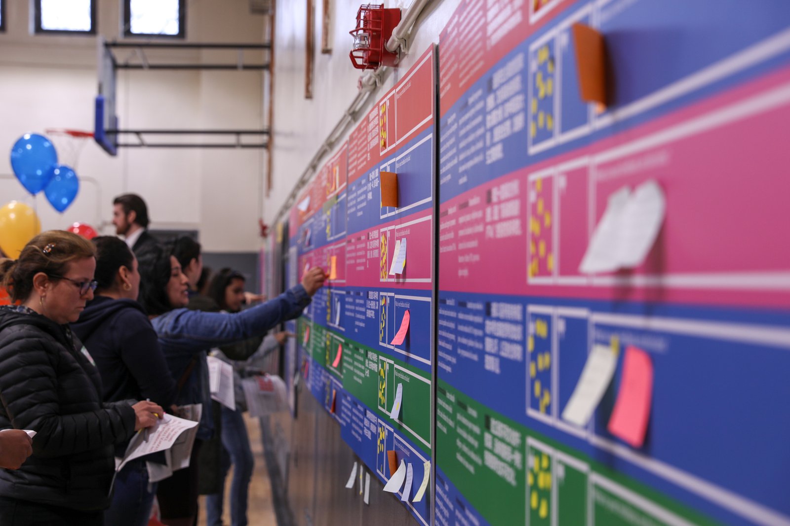 People interact with a colorful wall display covered in sticky notes and information panels.