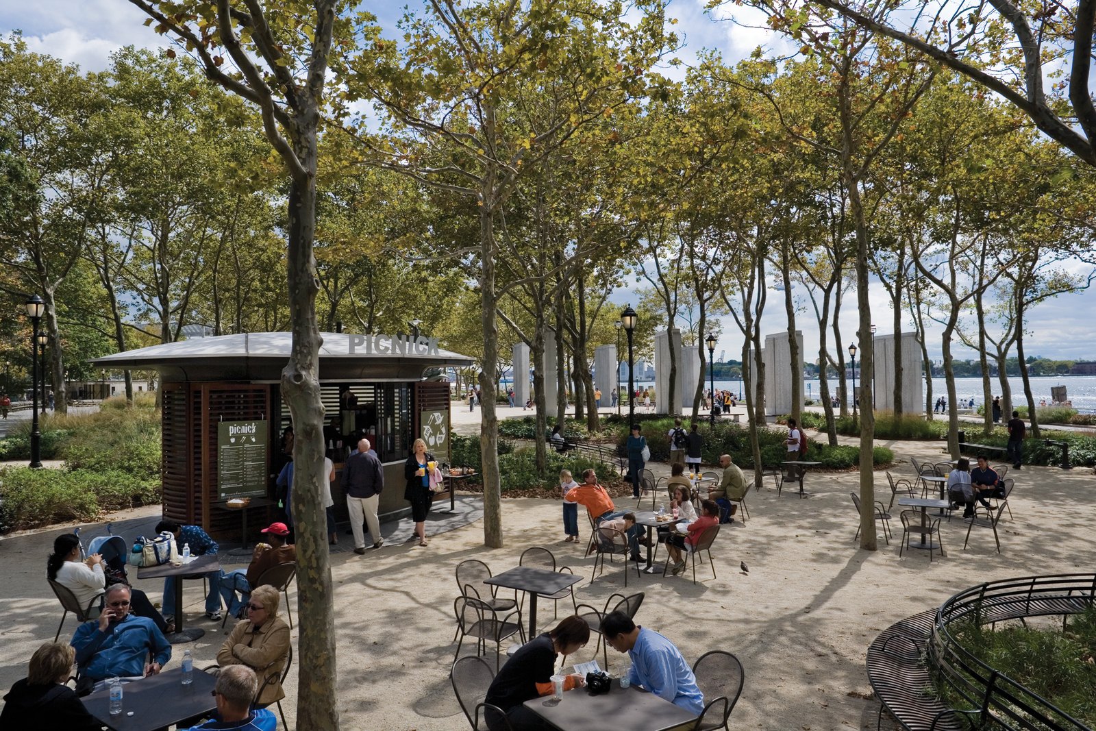 Visitors gather at outdoor tables near a food kiosk surrounded by trees and greenery.