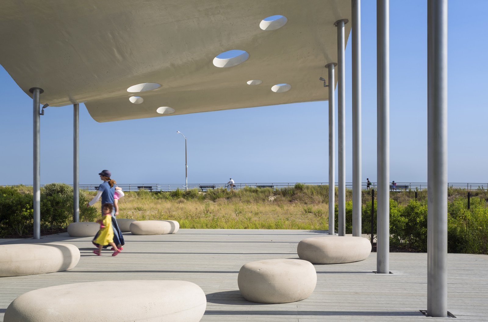 A shaded outdoor pavilion with perforated roof panels, stone seating, and views of grass dunes.