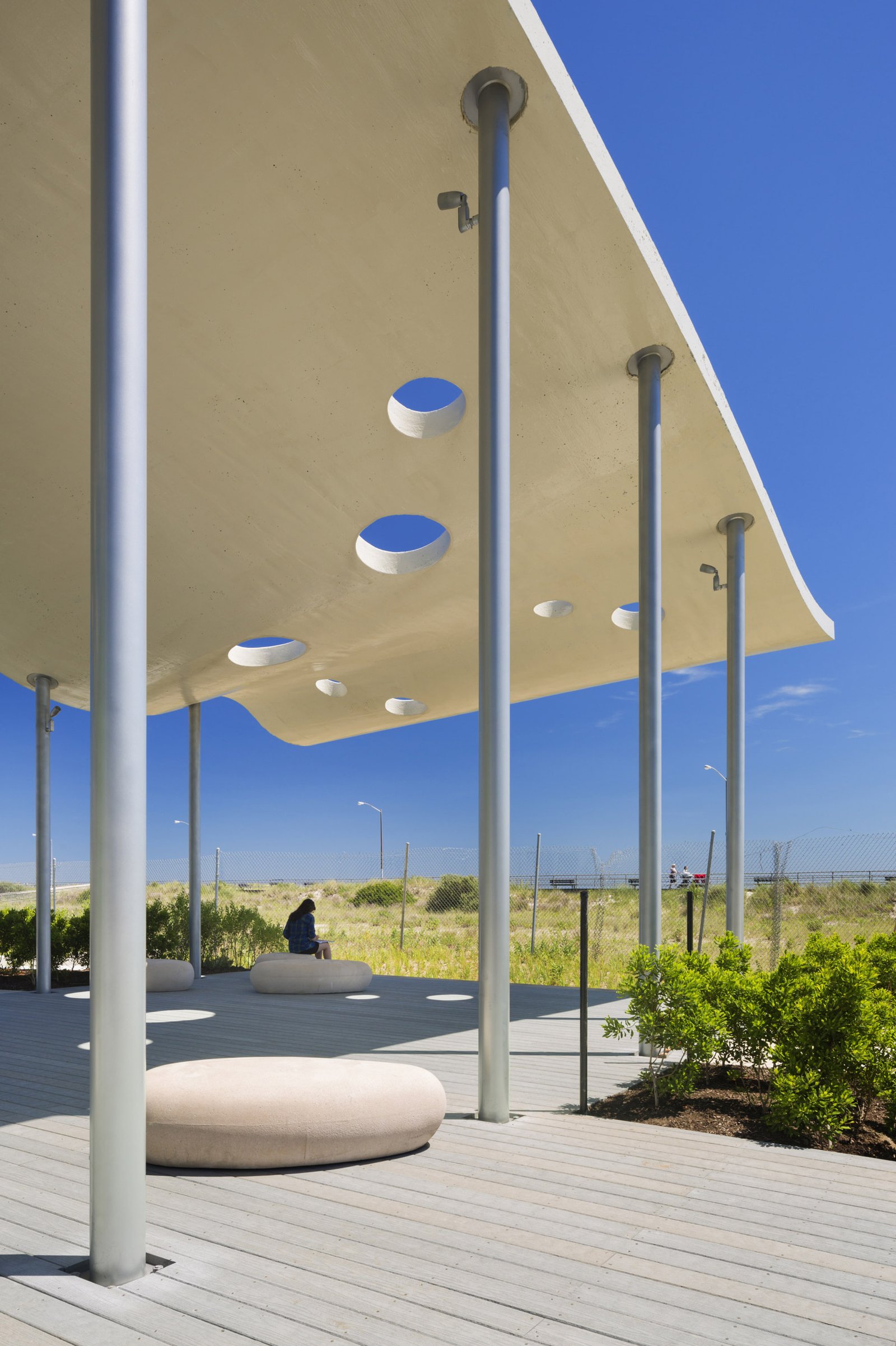 A pavilion with a perforated canopy, slender columns, and stone seating set in a grassy coastal park.