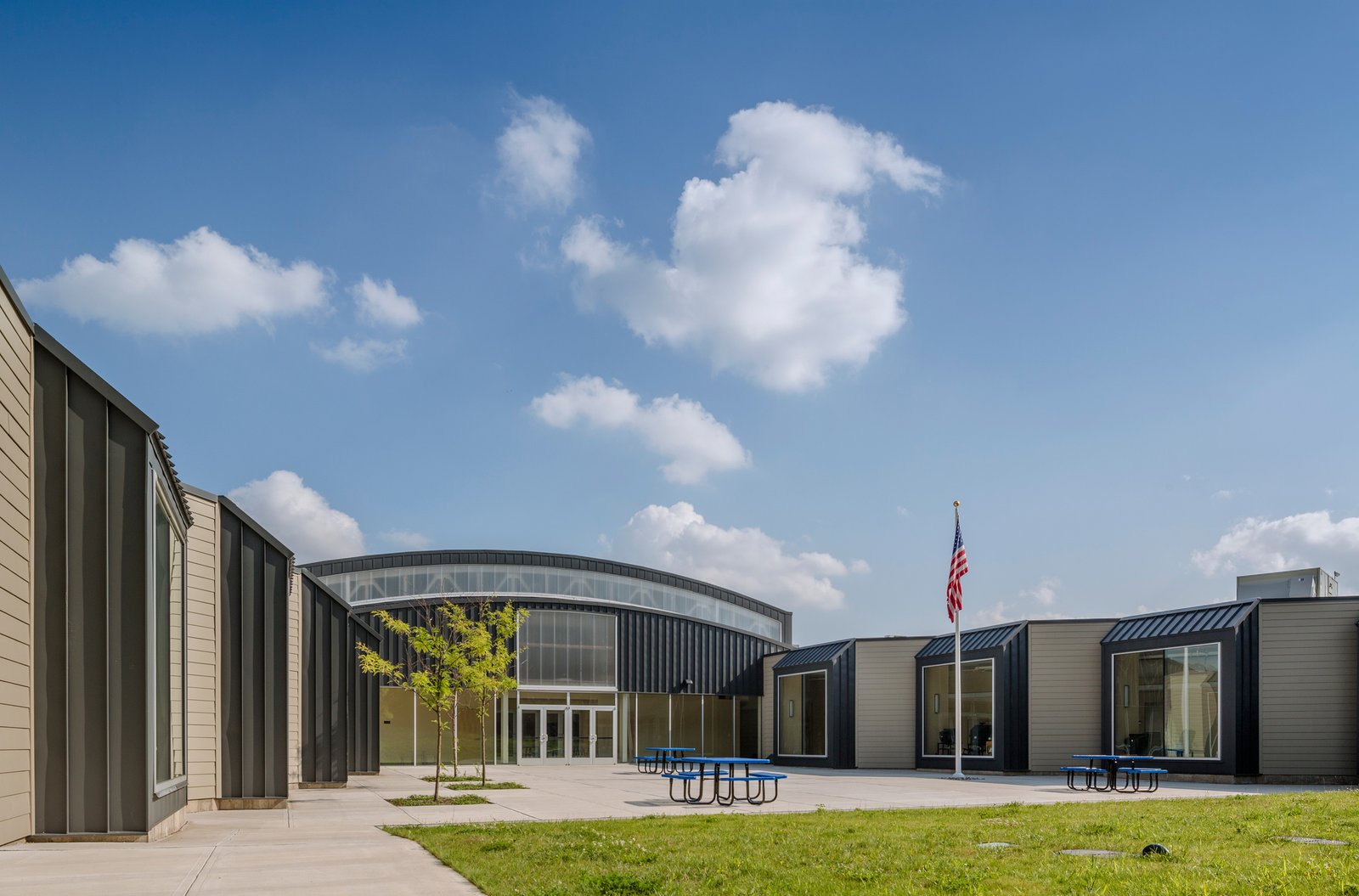 A school courtyard with modular buildings, large windows, and blue picnic tables under a bright sky.