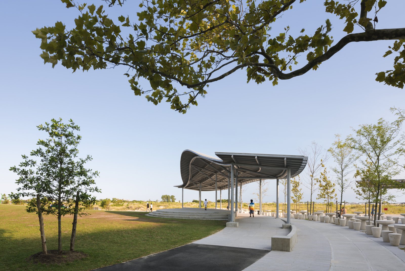 A curved canopy with metal roofing, slender columns, and a paved pathway in a grassy park.