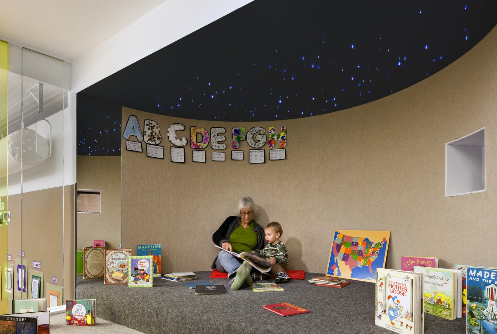 A reading nook with a dark ceiling dotted with small lights, books, and a caregiver reading to a child.