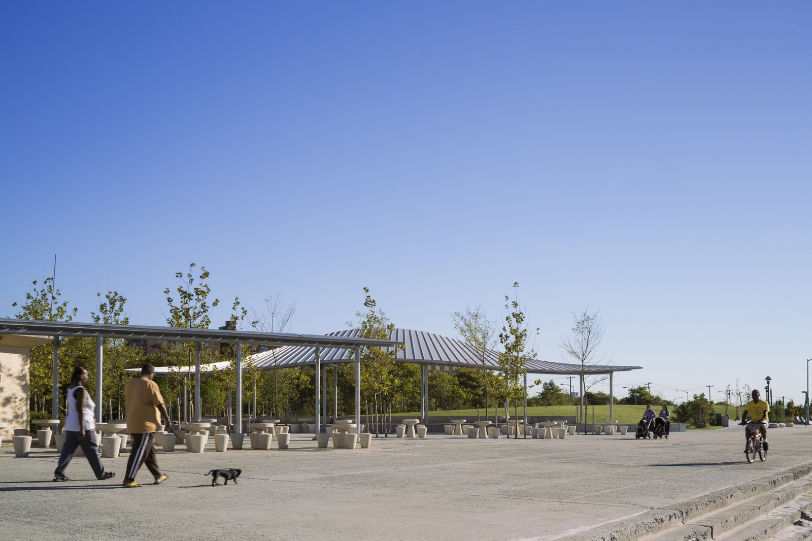 A pavilion with a metal roof, slender columns, and outdoor seating near a pedestrian walkway.