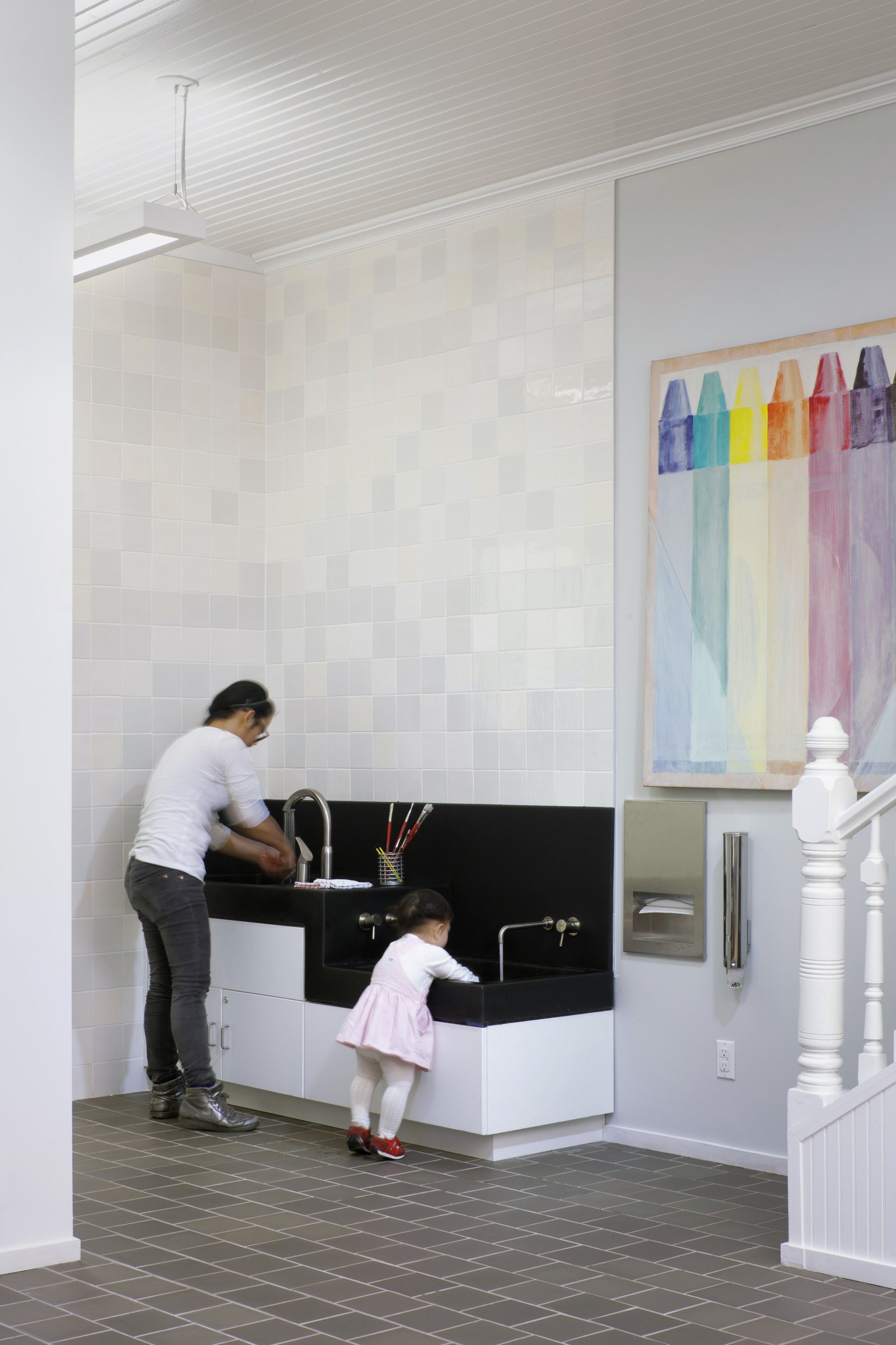 A child and an adult use a two-level black sink against a tiled wall, with a crayon painting nearby.