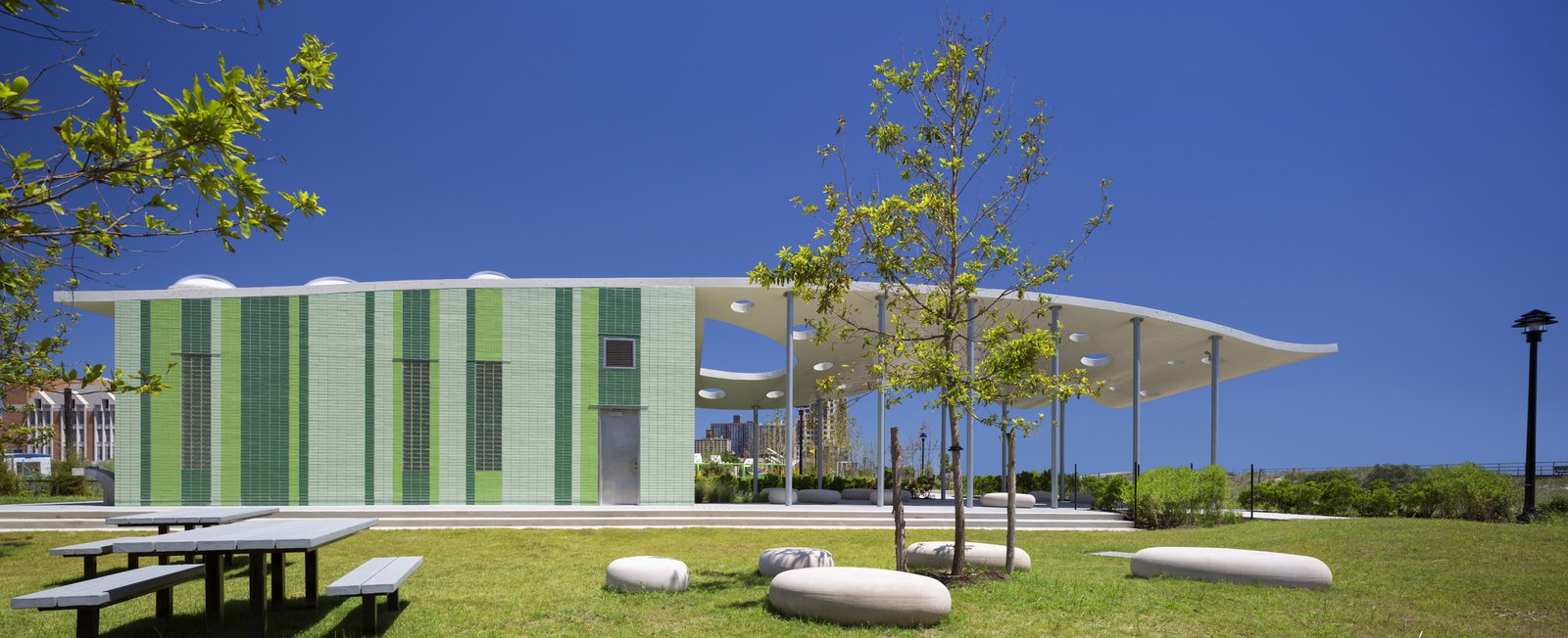 A comfort station with green glazed brick and a canopy, set in a grassy park with stone seating.