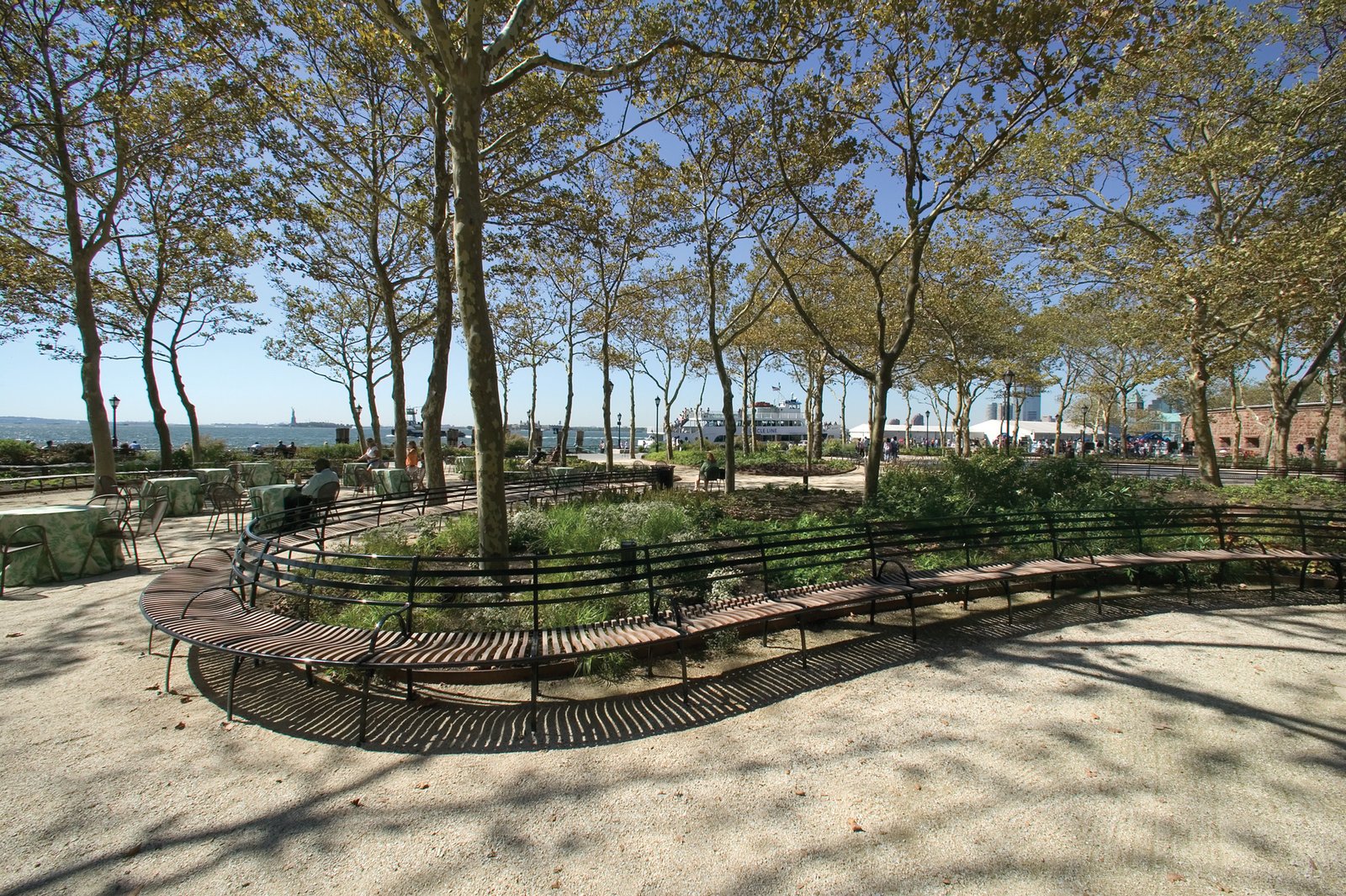 Curved wooden benches and tables under trees face the New York Harbor with clear views of the water.