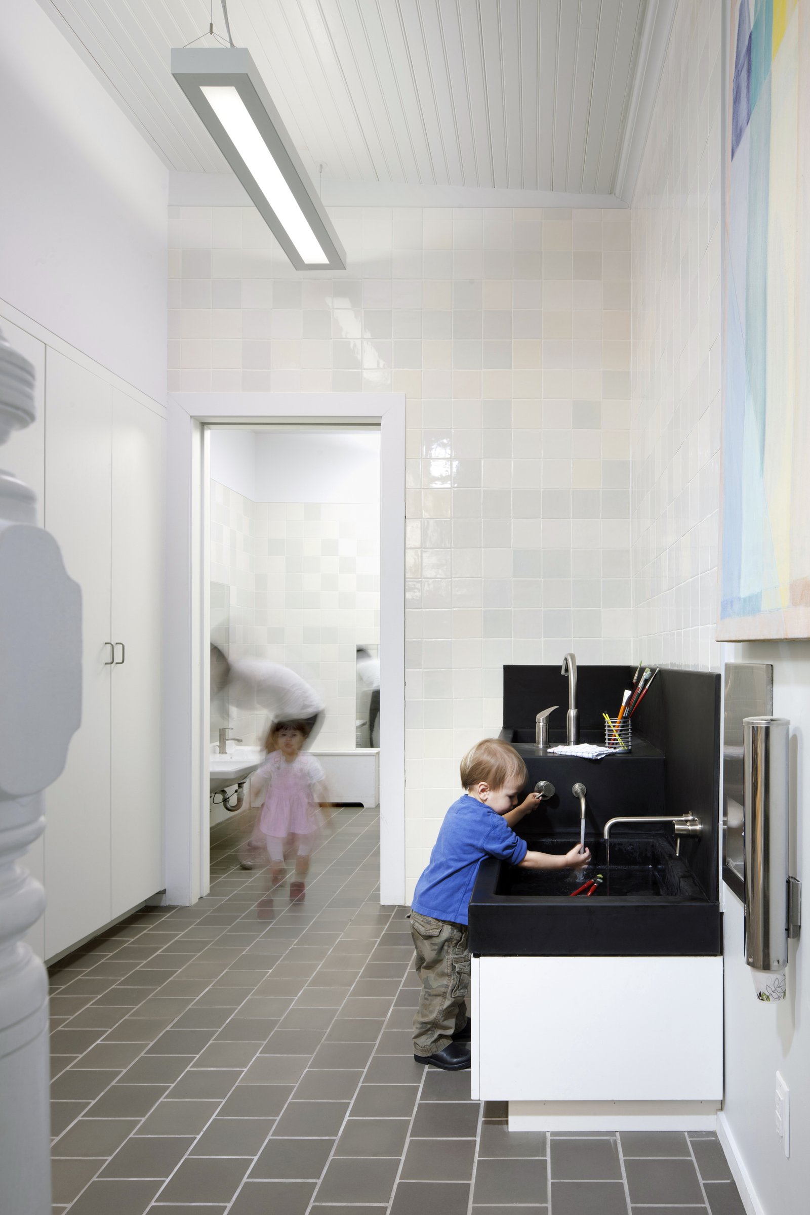 A child uses a black tiered sink while another child and an adult move in the background near a restroom.